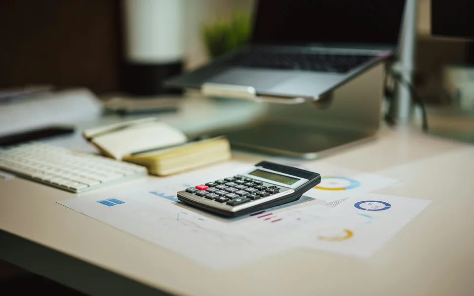 A calculator and financial papers beside a laptop.